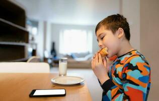 friska unge har frukost, Lycklig barn pojke använder sig av mobil telefon tittar på, läsning eller spelar spel en medan äter mat, skola unge croissant och mjölk innan gå till skola i de morgon- foto