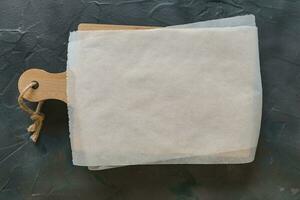 trä- skärande styrelse och vit pergament papper på mörk grå bakgrund. attrapp för recept, handla lista eller mat foto