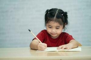 asiatisk bebis flicka bär en röd skjorta skriva anteckningar i anteckningsbok och läsning bok till studie uppkopplad på trä tabell skrivbord i levande rum på Hem. utbildning inlärning uppkopplad från Hem begrepp. foto