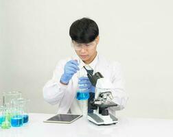 asiatisk manlig studerande forskare bär en läkares klänning i de labb ser på de kemist. orsakade förbi blandning reagens i vetenskaplig forskning laboratorier med testa rör och mikroskop på de tabell foto