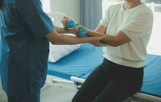 Lycklig senior kvinna håller på med övning på Hem med fysioterapeut. gammal pensionerad lady håller på med stretching vapen på Hem med de hjälp av en personlig tränare under en rehabilitering session. foto