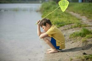 en liten pojke med en fjäril netto sitter nära de sjö och utseende på de vatten. foto