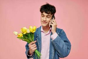 ung kille i en denim jacka med en bukett av blommor talande på de telefon rosa bakgrund foto