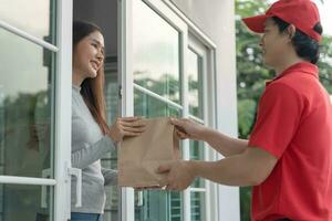 Lycklig leende asiatisk kvinna mottar papper väska paket av mat från kurir främre hus. leverans man skicka leverera uttrycka. uppkopplad handla, papper behållare, hämtmat, brevbärare, leverans service, paket foto