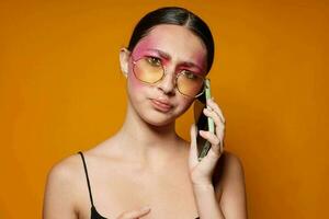 sexig brunett kvinna med telefon rosa ansikte smink Framställ attraktiv se glasögon gul bakgrund oförändrad foto