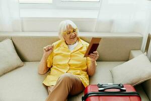 Lycklig senior kvinna med pass och resa biljett packade en röd resväska, semester och hälsa vård. leende gammal kvinna lyckligt sitter på de soffa innan de resa. foto