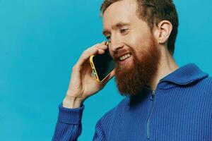 porträtt av en man med en telefon i hans händer gör ser på den och talande på de telefon, på en blå bakgrund. kommunicerar uppkopplad social media, livsstil foto