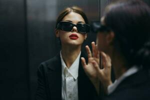 eleganta kvinna i en svart jacka och solglasögon Framställ i ett hiss hiss nära de spegel, mode modell, mörk filmiska ljus och Färg matris, glamour årgång foto