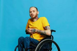 en man i en rullstol leende utseende på de kamera i en t-shirt med tatueringar på hans vapen sitter på en blå studio bakgrund, en full liv, en verklig person foto