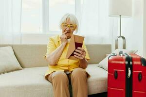 Lycklig senior kvinna med pass och resa biljett packade en röd resväska, semester och hälsa vård. leende gammal kvinna glatt Sammanträde på de soffa innan de resa Uppfostrad henne händer upp i glädje. foto