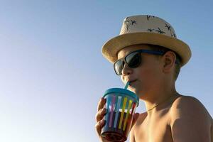 Lycklig pojke i hatt och solglasögon dricka juice på de strand foto