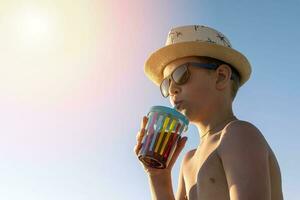 Lycklig pojke i hatt och solglasögon dricka juice på de strand foto