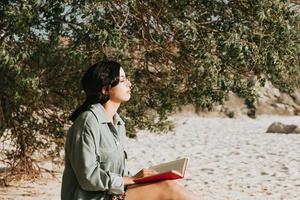 ung marockansk kvinna i moderna kläder som sitter på stranden och läser en bok foto