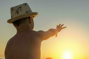 Lycklig pojke sitter på de strand på solnedgång , sträckor hans hand för de Sol foto