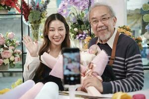 e-handel företag, asiatisk äldre man och dotter blomsterhandlare visar blommig arrangemang och samtal till kamera via uppkopplad live streaming webbkamera Ansökan i en ljus blomma affär, skön blommar Lagra. foto
