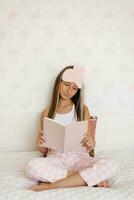 Tonårs flicka är Sammanträde på de säng i henne pyjamas i de morgon- och läsning en bok. mysigt helgen på Hem foto