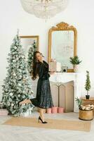 porträtt av en skön ung leende kvinna i en svart klänning med paljetter, innehav en gåva i henne händer. fira jul i de levande rum förbi de jul träd och öppen spis foto