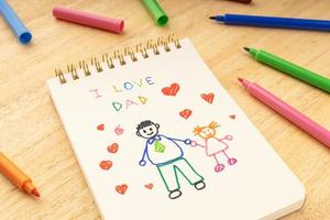 fäder dag koncept anteckningsbok med ritning av en far med en flicka och markör pennor på ett träbord foto