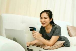 asiatisk kvinna som håller ett kreditkort för att shoppa online medan du sitter på soffan medan du är på semester hemma. foto