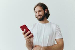 en man med en skägg i vit t-shirt och jeans utseende på hans telefon vända genom ett uppkopplad social media utfodra med hörlurar i hans öron lyssnande till en röst meddelande på en vit bakgrund och leende foto