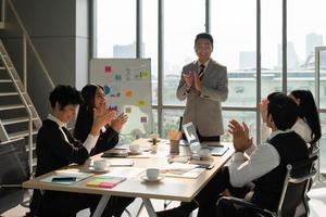 medelålders asiatisk chef som klappar händer med multiracialt team för framgång för nytt projekt i kontorsmötesrum foto