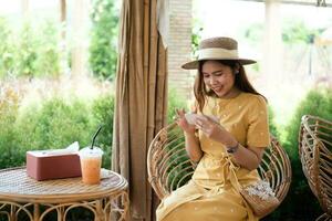 asiatisk kvinna med skön leende läsning Bra Nyheter på mobil telefon under resten i kaffe affär, Lycklig kvinna tittar på henne foton på cell telefon medan avkopplande i Kafé under fri tid
