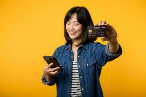 porträtt skön ung asiatisk kvinna Lycklig leende klädd i denim jacka som visar smartphone och kreditera kort isolera på gul studio bakgrund. handla uppkopplad smartphone Ansökan begrepp foto