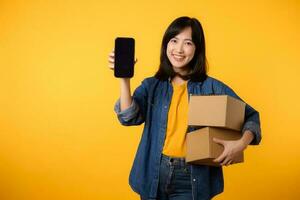 porträtt asiatisk ung kvinna bär gul t-shirt och denim skjorta innehav mobil telefon och paket låda isolerat på gul studio bakgrund, leverans kurir och frakt service begrepp. foto