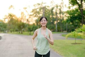 passa asiatisk ung kvinna joggning i parkera leende Lycklig löpning och njuter en friska utomhus- livsstil. kvinna joggare. kondition löpare flicka i offentlig parkera. friska livsstil och wellness varelse begrepp foto