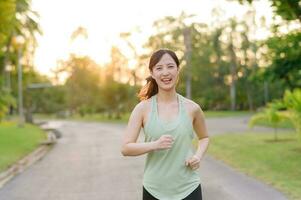 passa asiatisk ung kvinna joggning i parkera leende Lycklig löpning och njuter en friska utomhus- livsstil. kvinna joggare. kondition löpare flicka i offentlig parkera. friska livsstil och wellness varelse begrepp foto