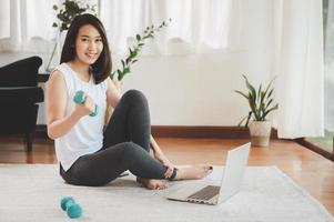 hälsosam vacker asiatisk kvinnasammanträde på golvinnehavhanteln som använder bärbara datorn hemma i vardagsrum redo för online-träning foto