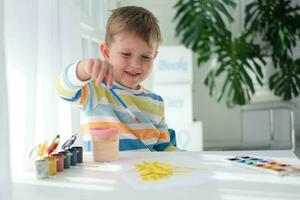 Lycklig unge målning med borsta och färgrik färger. en liten pojke drar en bild med målarfärger på en ark av papper. de barn är inlärning till dra. Hem kreativitet. foto