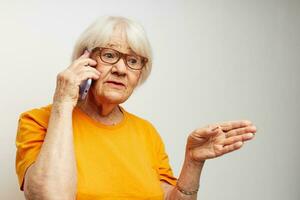 porträtt av ett gammal vänlig kvinna i tillfällig t-shirt glasögon talande på de telefon ljus bakgrund foto