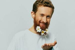 porträtt av en rolig man i en vit t-shirt med blommor daisy i hans skägg på en vit isolerat bakgrund, kopia plats. Semester begrepp och grattis. foto