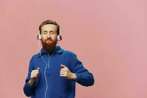 porträtt av en rödhårig man bär hörlurar leende och dans, lyssnande till musik på en rosa bakgrund. en hipster med en skägg. foto
