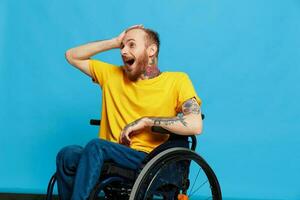 en man i en rullstol överraskad och öppen mun i en t-shirt med tatueringar på hans vapen sitter på en blå studio bakgrund, en full liv, en verklig person foto