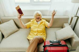 Lycklig senior kvinna med pass och resa biljett packade en röd resväska, semester och hälsa vård. leende gammal kvinna glatt Sammanträde på de soffa innan de resa Uppfostrad henne händer upp i glädje. foto
