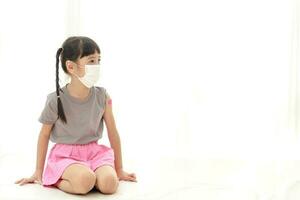 söt liten asiatisk flicka med pigtails ha på sig en vit mask Sammanträde på en vit golv. begrepp av vaccination för barn till förhindra coronavirus. kopia Plats foto