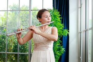 asiatisk kvinna bär en grädde färgad klänning hon är spelar de flöjt, ett internationell vind instrument. spela klassisk sånger. utför konserter spelar klassisk musik i en band. musik inlärning foto