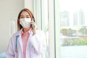 skön asiatisk kvinna läkare bär en vit mask håll en smartphone och prata till de patient. modern kommunikation teknologi begrepp, patient vård genom smartphone. kopia Plats foto