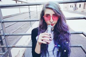 glad vacker tonåring med rosa solglasögon dricker och njuter av en rosa dryck som sitter på stadsmarken foto