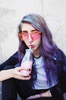 glad vacker tonåring med rosa solglasögon dricker och njuter av en rosa dryck som sitter på stadsmarken foto