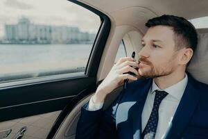 allvarlig verkställande i blå kostym funderar Viktig beslut, väger risker och vinster, innehar telefon mot haka i limousine. foto
