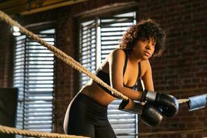 kvinna kämpe flicka kraft. afrikansk amerikan kvinna kämpe med boxning handskar stående på boxning ringa lutande på tågvirke väntar och vilar. stark kraftfull flicka. styrka passa kropp träna Träning. foto