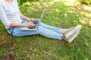 kvinna ben på grönt gräs gräsmatta i stadsparken, händer som arbetar på bärbar dator. frilansande affärsidé foto