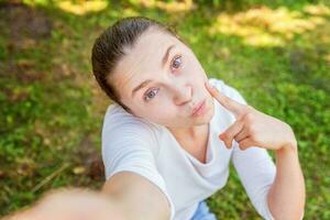 ung flicka ta selfie från händer med telefon som visar rolig ansikte Sammanträde på parkera eller trädgård bakgrund foto