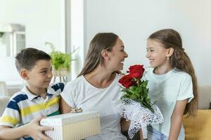 glad liten flicka med bukett av ro blommor och yngst bror med gåva låda leende och gratulera Lycklig mamma på mor dag på Hem. Lycklig mödrar dag foto