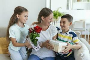 glad liten flicka med gåva låda och yngst bror med bukett av ro blommor leende och gratulera Lycklig mamma på mor dag på Hem. Lycklig mödrar dag foto