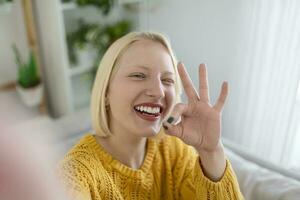 busig ung kvinna tar selfie på Hem. skön blond kvinna ok tecken med hand blinkning med ett öga och med henne tunga ut foto