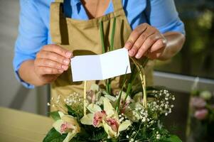 närbild kvinna blomsterhandlare innehav vit tom Grattis kort över en söt blomma arrangemang i en korg- korg foto